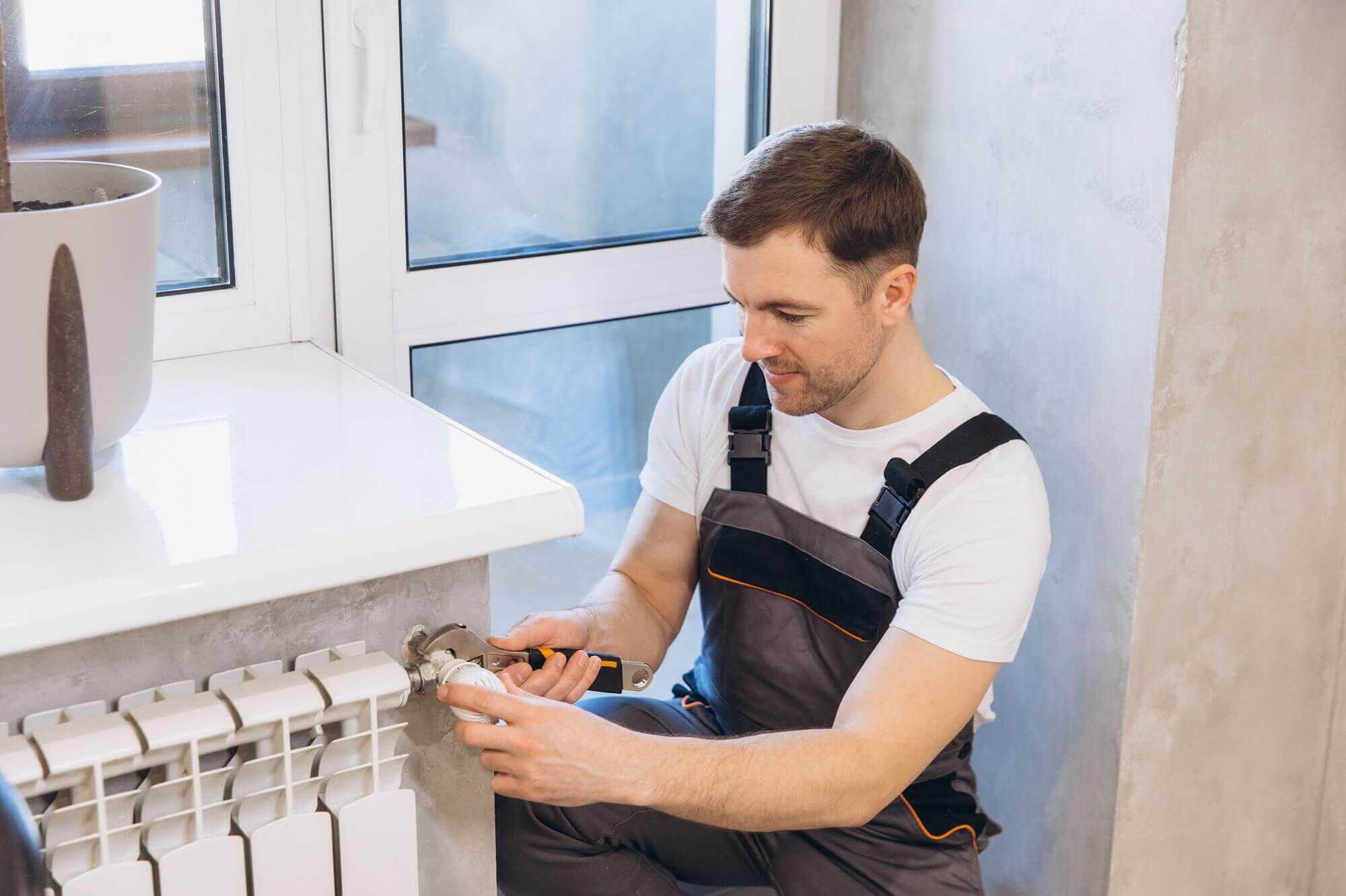 Na obrazku widać hydraulika, który naprawia lub montuje instalację przy grzejniku w pomieszczeniu mieszkalnym. Mężczyzna używa klucza, aby dokręcić zawór lub element instalacji grzewczej. Pracuje przy oknie, a obok znajduje się parapet z doniczką.