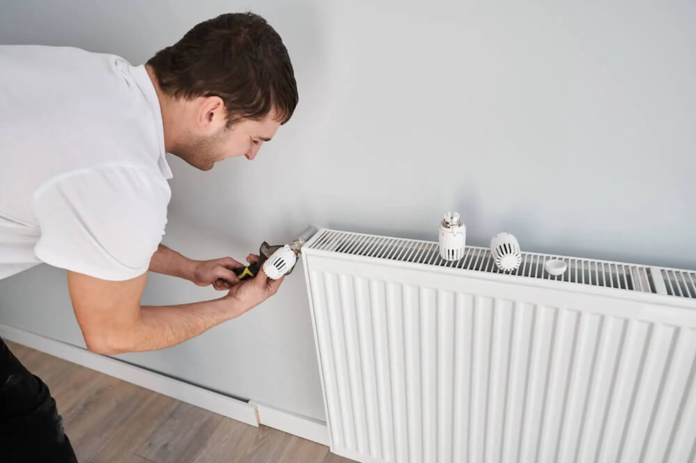 Na obrazku widać mężczyznę, który montuje lub naprawia zawór przy grzejniku. Używa narzędzia, aby dokręcić element instalacji grzewczej, skupiając się na precyzyjnej pracy. Obok na grzejniku leżą części zaworu, co wskazuje na trwającą wymianę lub serwis.