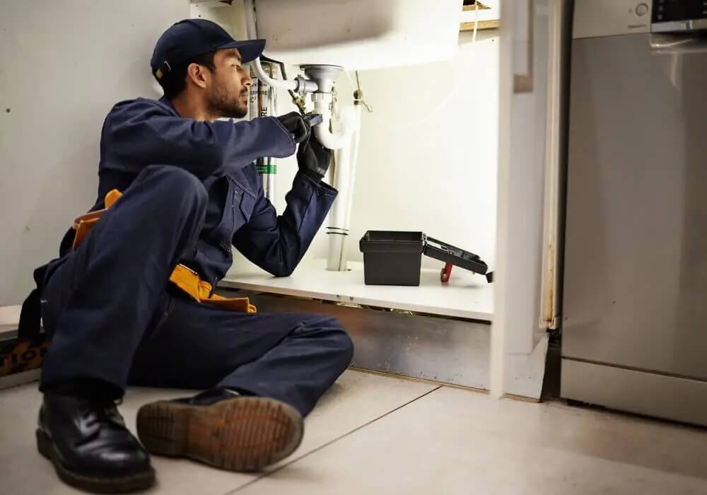 Na zdjęciu przedstawiono hydraulika wykonującego naprawę lub montaż instalacji pod zlewem w kuchni. Fachowiec sprawdza elementy odpływu i syfonu, a obok widoczne są skrzynka z narzędziami oraz sprzęt niezbędny do wykonania prac hydraulicznych.
