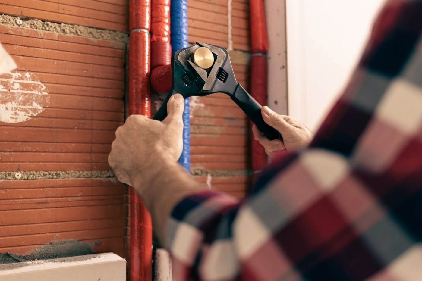 Na zdjęciu widać hydraulika wykonującego prace przy instalacji wodnej. Za pomocą specjalistycznego klucza dokonuje regulacji lub naprawy rur umieszczonych w ścianie. Fotografia ilustruje profesjonalne usługi hydrauliczne związane z diagnostyką, usuwaniem awarii oraz naprawą instalacji wodnych, w tym wykrywaniem i usuwaniem wycieków wody.