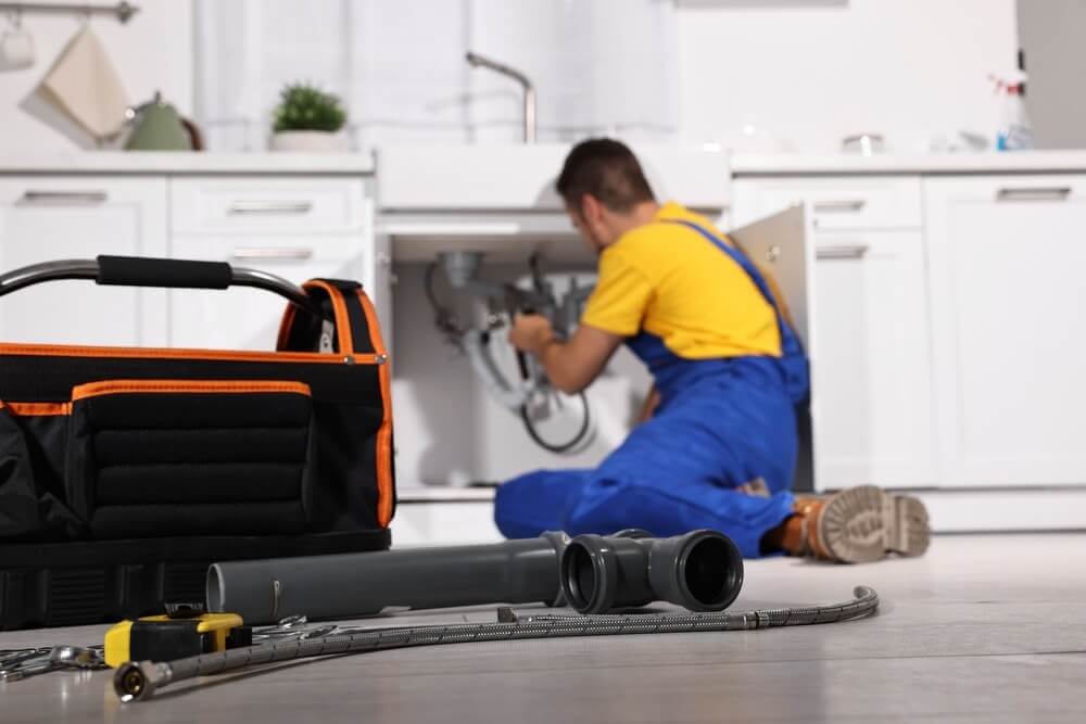 Na zdjęciu przedstawiono hydraulika wykonującego prace instalacyjne pod zlewem w nowoczesnej kuchni. Na pierwszym planie widoczne są narzędzia, elementy instalacji hydraulicznej oraz torba serwisowa, przygotowane do montażu lub naprawy.