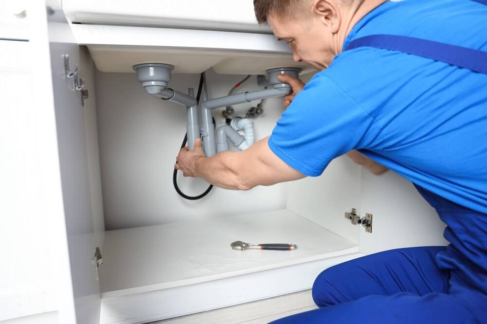 Na zdjęciu przedstawiono hydraulika wykonującego montaż lub naprawę instalacji odpływowej pod zlewem. Fachowiec dopasowuje i łączy elementy syfonu oraz rur kanalizacyjnych znajdujących się wewnątrz szafki kuchennej. Na dnie szafki leży klucz wykorzystywany podczas prac montażowych. Fotografia prezentuje typowe prace hydrauliczne związane z instalacją, wymianą lub serwisem syfonu oraz podłączeniem odpływu zlewozmywaka.